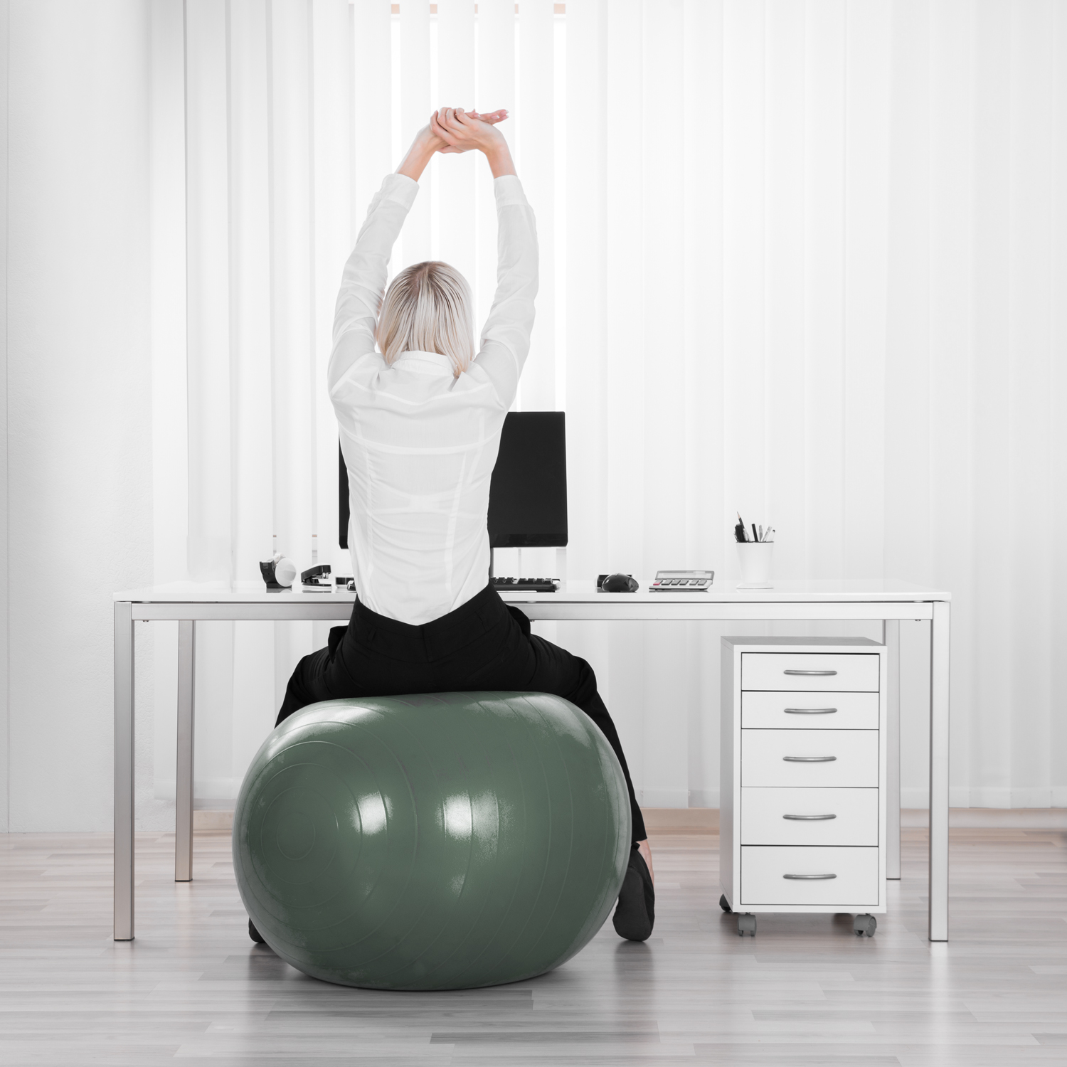 Rückansicht einer Frau, die an einem Schreibtisch auf einem Gymnastikball sitzt und mit nach oben gestreckten Armen ihren Rücken dehnt – ein Beispiel für Betriebliche Gesundheitsförderung am Arbeitsplatz.