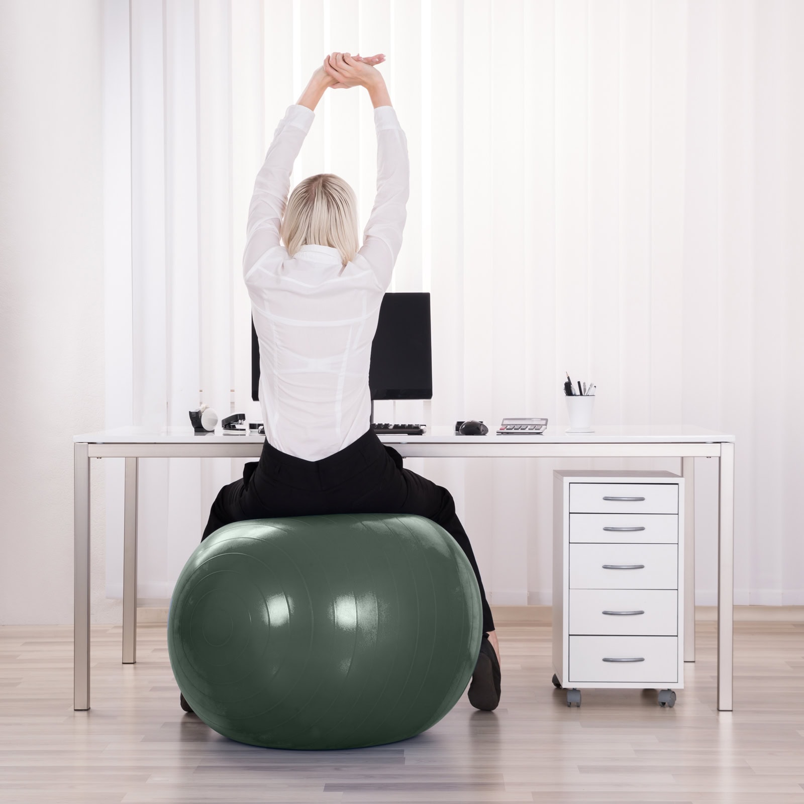 Rückansicht einer Frau, die an einem Schreibtisch auf einem Gymnastikball sitzt und mit nach oben gestreckten Armen ihren Rücken dehnt – ein Beispiel für Betriebliche Gesundheitsförderung am Arbeitsplatz.